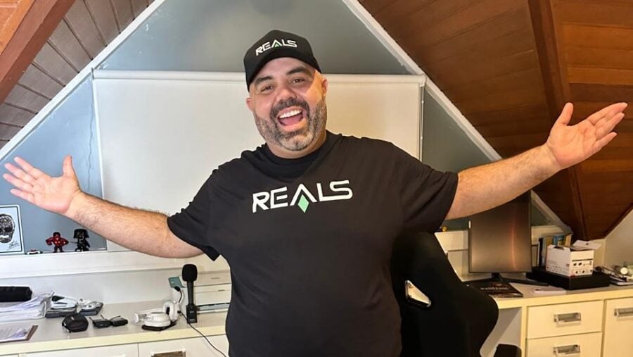 (Edu Primitivo Fecha com Casa de Apostas Realsbet). Homem sorridente, de barba e boné preto com a marca Reals, veste camiseta da mesma empresa e posa com os braços abertos em um escritório doméstico com teto inclinado, escrivaninha branca e equipamentos eletrônicos ao fundo.