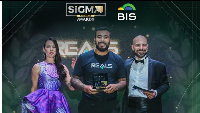 Rafael Borges, usando camiseta com o logo da Reals, segura um troféu no centro do palco entre uma mulher de vestido roxo e um homem de smoking, durante a cerimônia de premiação SiGMA Americas Awards 2023, com os logos do evento e do BIS ao fundo.